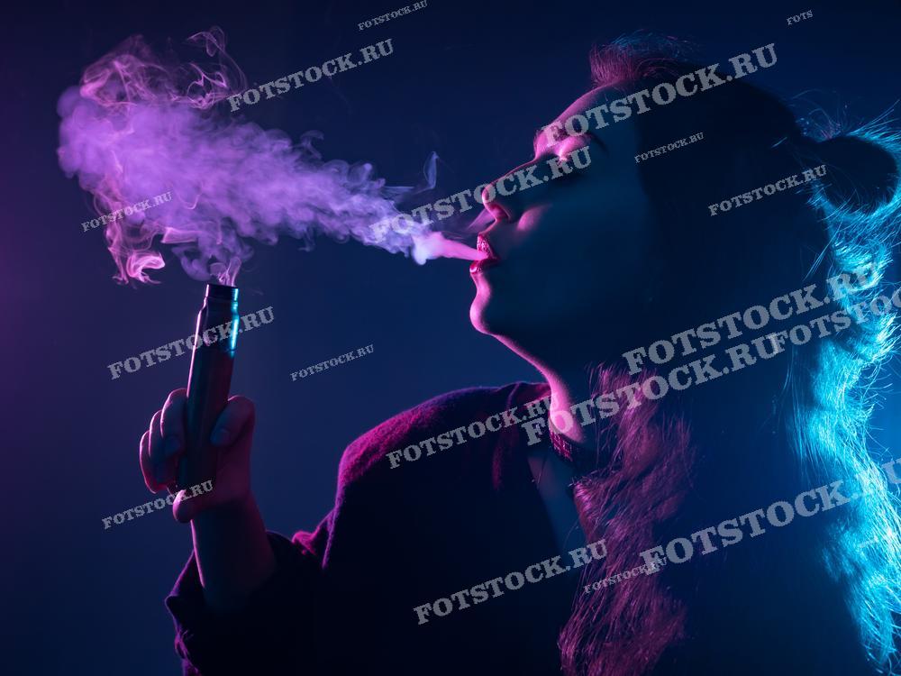 На фотографии изображена девушка с длинными волосами, держащая вейп-устройство и выпускающая густое облако дыма. Световое освещение создает атмосферу загадочности, подчеркивая детали фигуры и дыма.
